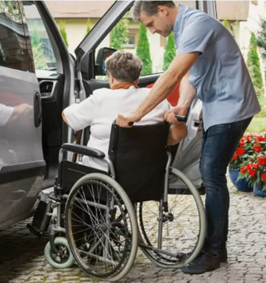 A man helping an elderly person in a wheelchair get into the back of a car.